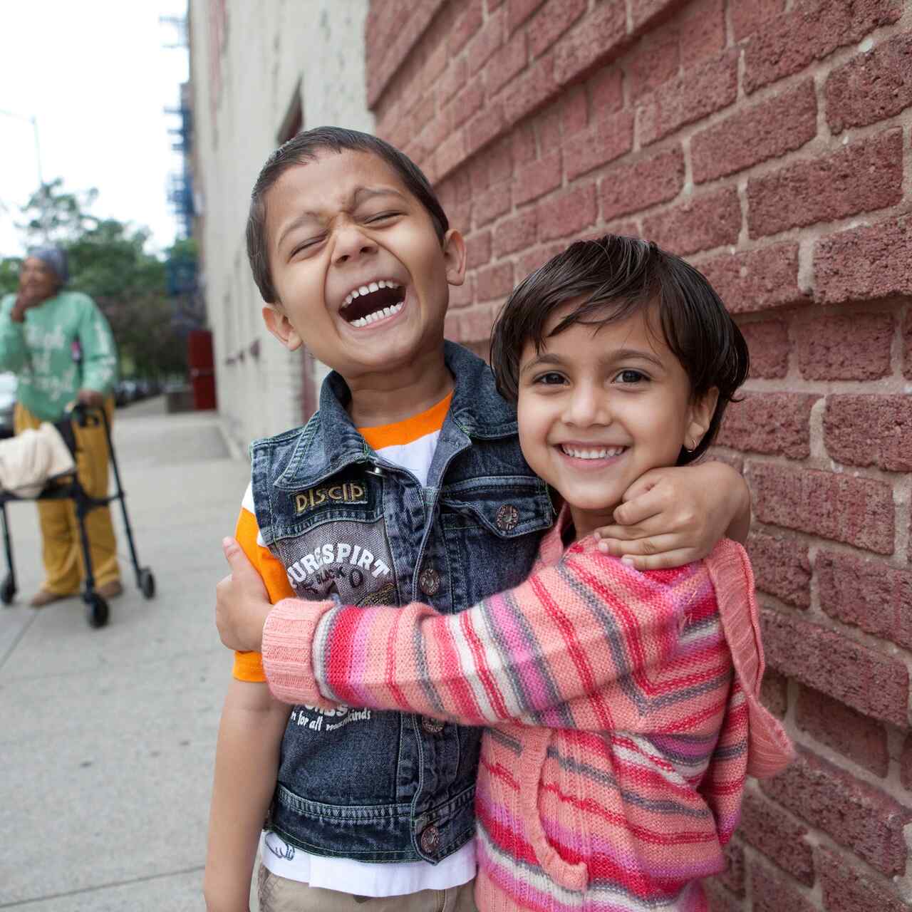 A young boy and girl stand outside in front of a brick wall, laughing and embracing.