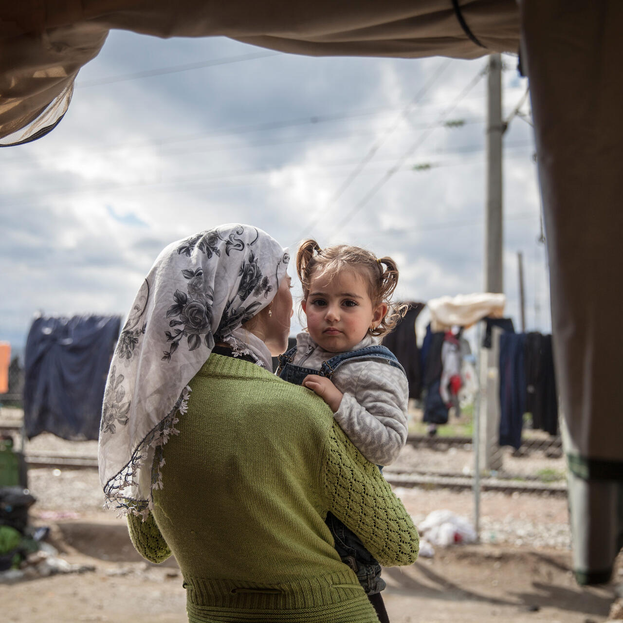 A mother holds her toddler daughter and looks out from their tent on a cloudy day in a refugee camp in Idomeni, Greece.