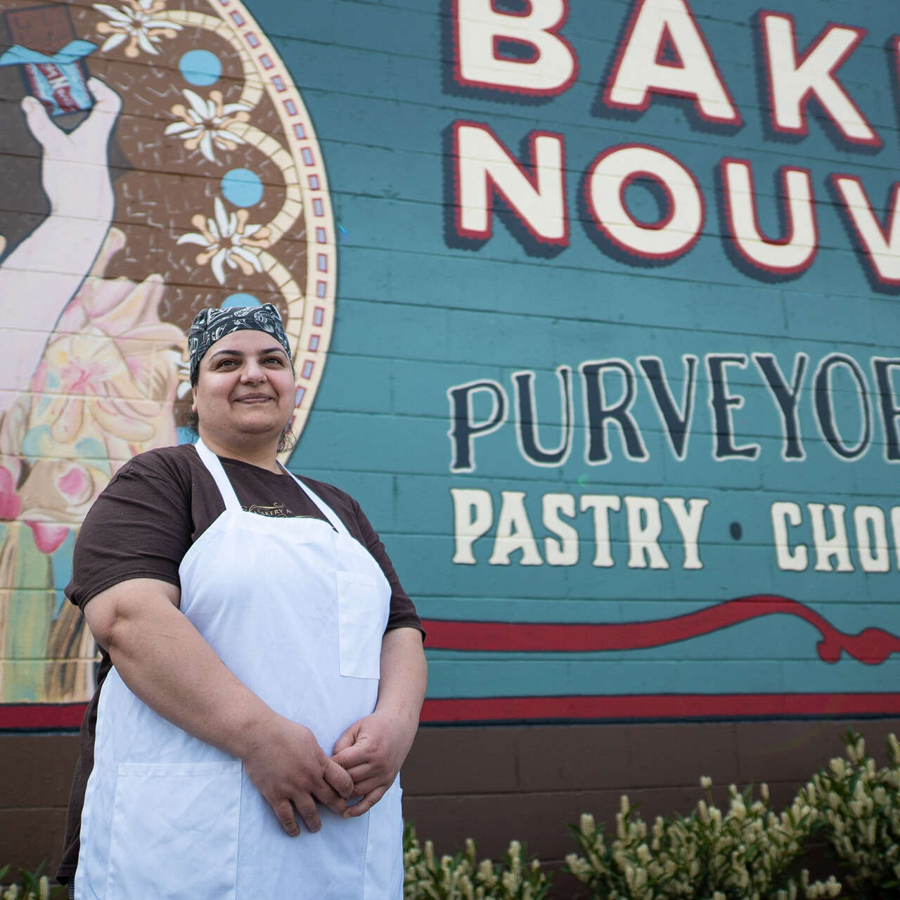 A woman stands by a mural on the side of a bakery and poses for a photo.