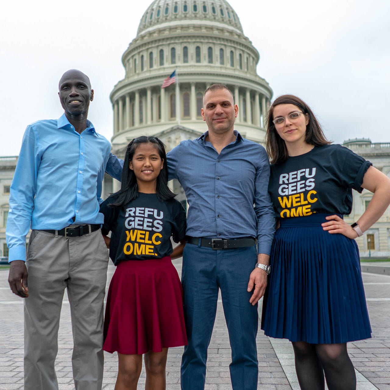 Four people pose for a picture in front of a capital building; two wear shorts with text reading "refugees welcome".