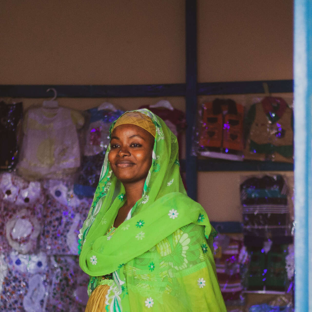 A woman, wearing a vibrant green headscarf and yellow dress, smiles in a shop in Cameroon.