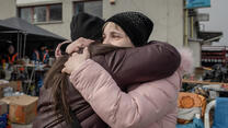 Two women wearing hats and winter coats hug outside.