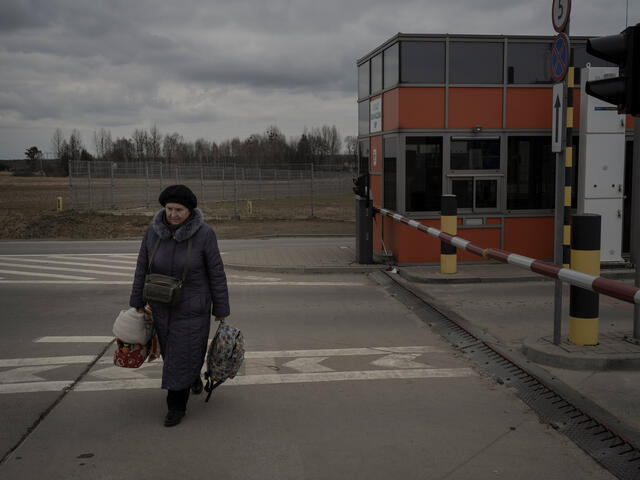 A woman carrying suitecases and wearing a winter hat and coat, walks across a parking lot. 