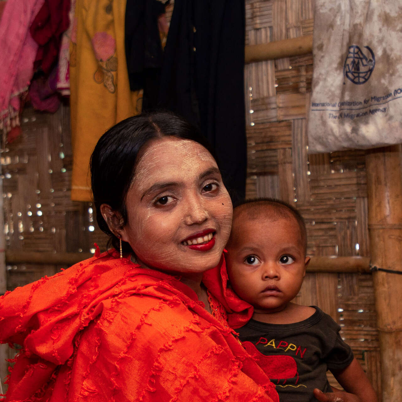 A mother, wearing a bright orange shirt, holds her young child and poses for a photo inside their home in Bangladesh.