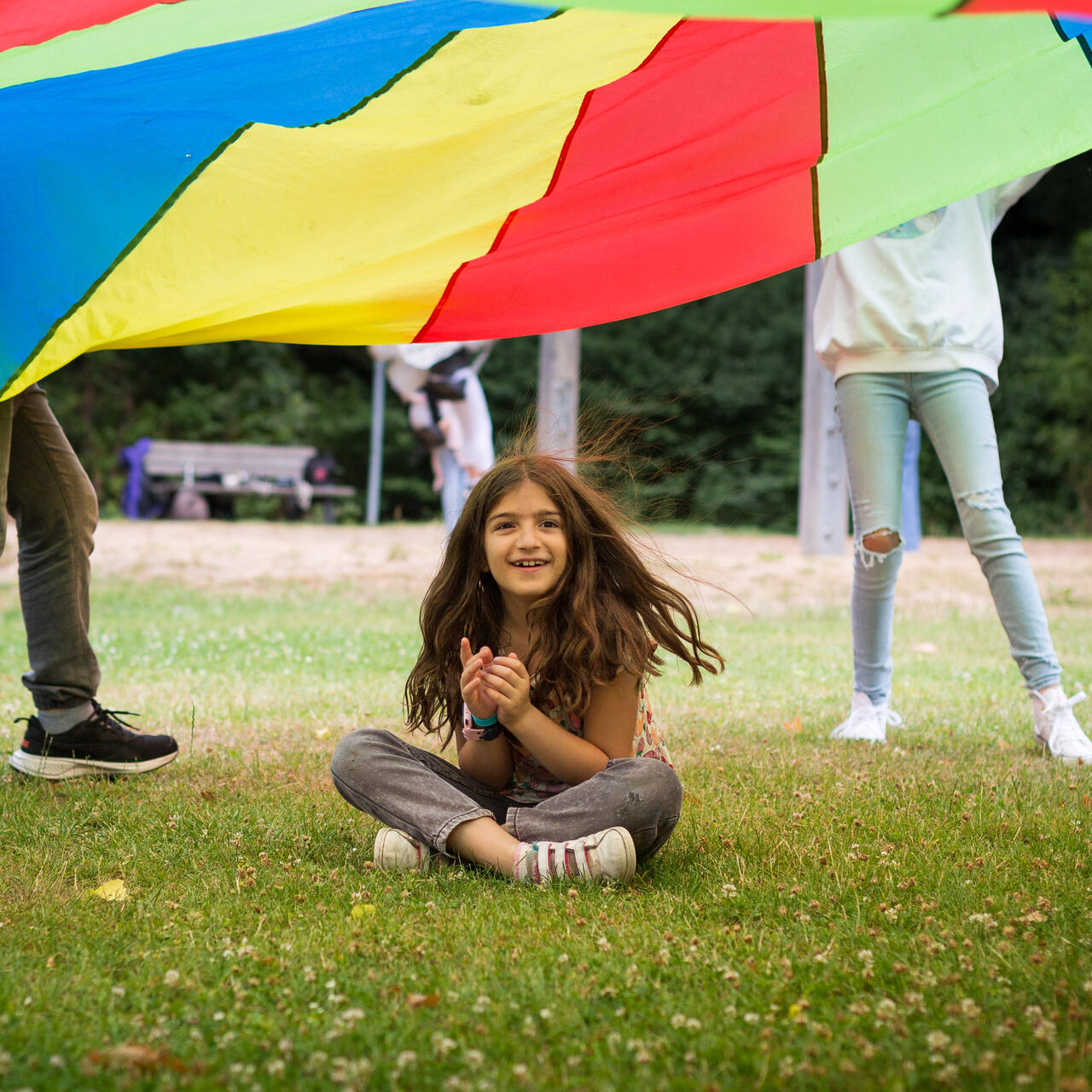 A girl sits under a colorful parachute while completing a group activity outdoors.