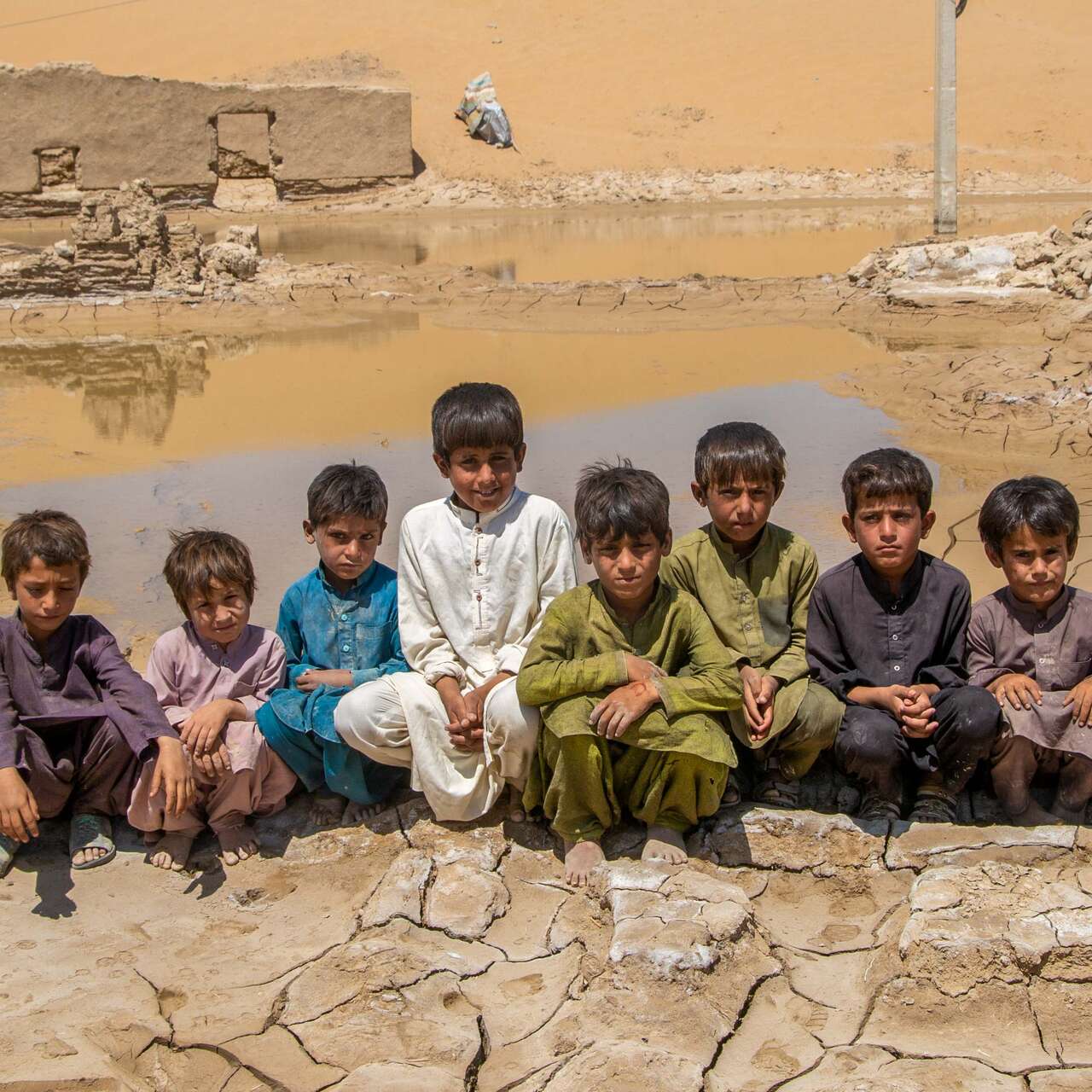 8 boys sit together outside in Pakistan, in front of a landscape devastated by flooding.