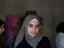 A young woman smiles at the camera in Lebanon. Behind her, other young women speak to an IRC staff member.