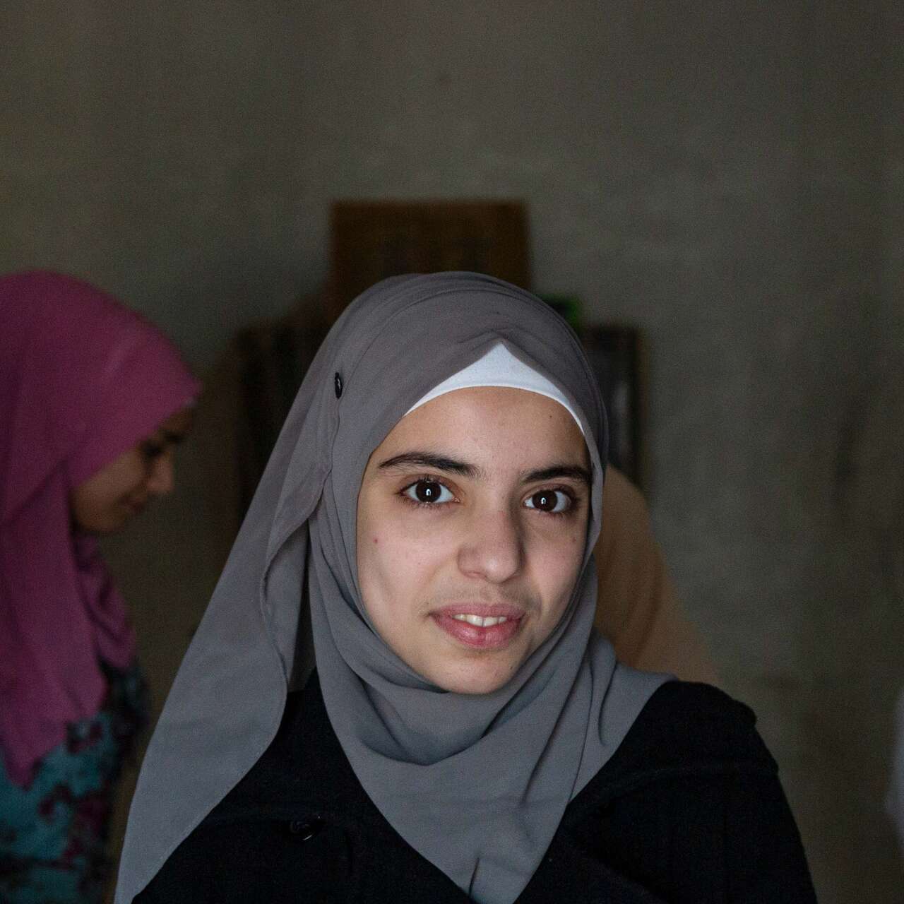 A young woman smiles at the camera in Lebanon. Behind her, other young women speak to an IRC staff member.