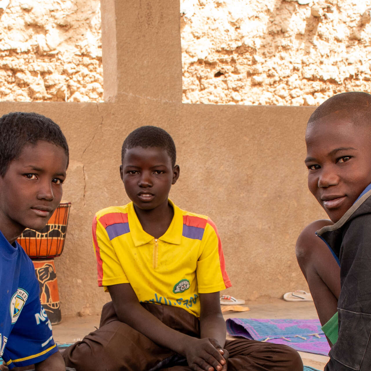 Three boys sit on the floor together in conversation. They look towards the camera.