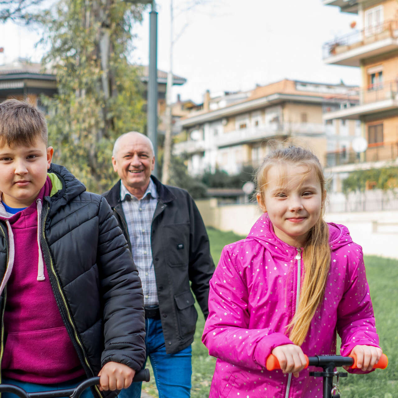 Two children ride scooters while their grandfather walks behind them.