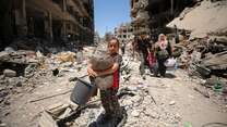 A girl holds belongings as she walks through rubble between destroyed buildings while people follow behind her in Gaza.