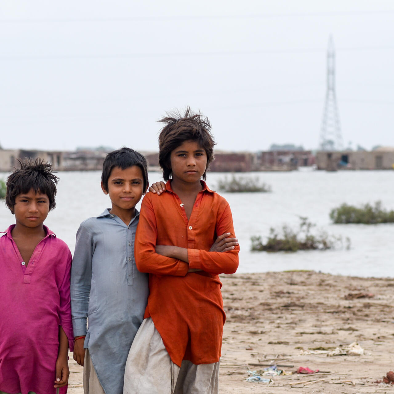 Three children pose for a photo in front of a community impacted by flooding.