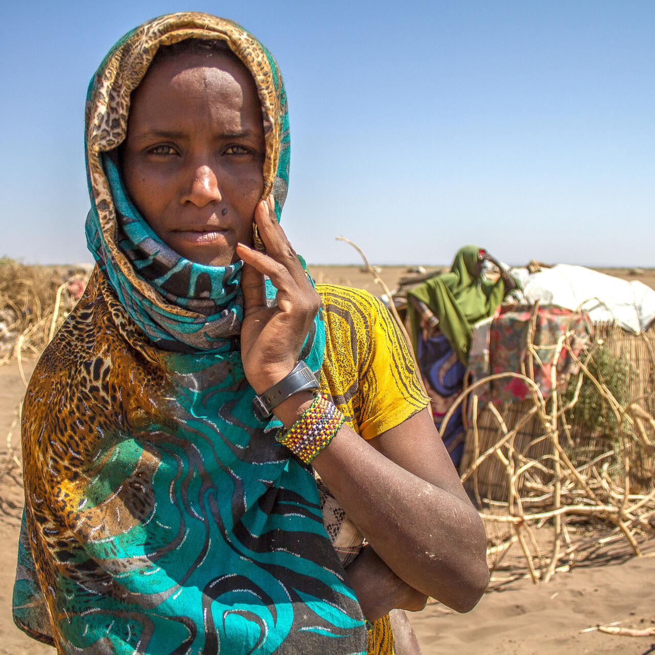 Deqa Omer, 32, stands among makeshift tents in a parched landscape in Ethiopia during a drought, her fingers resting on her cheek as she looks at the camera.