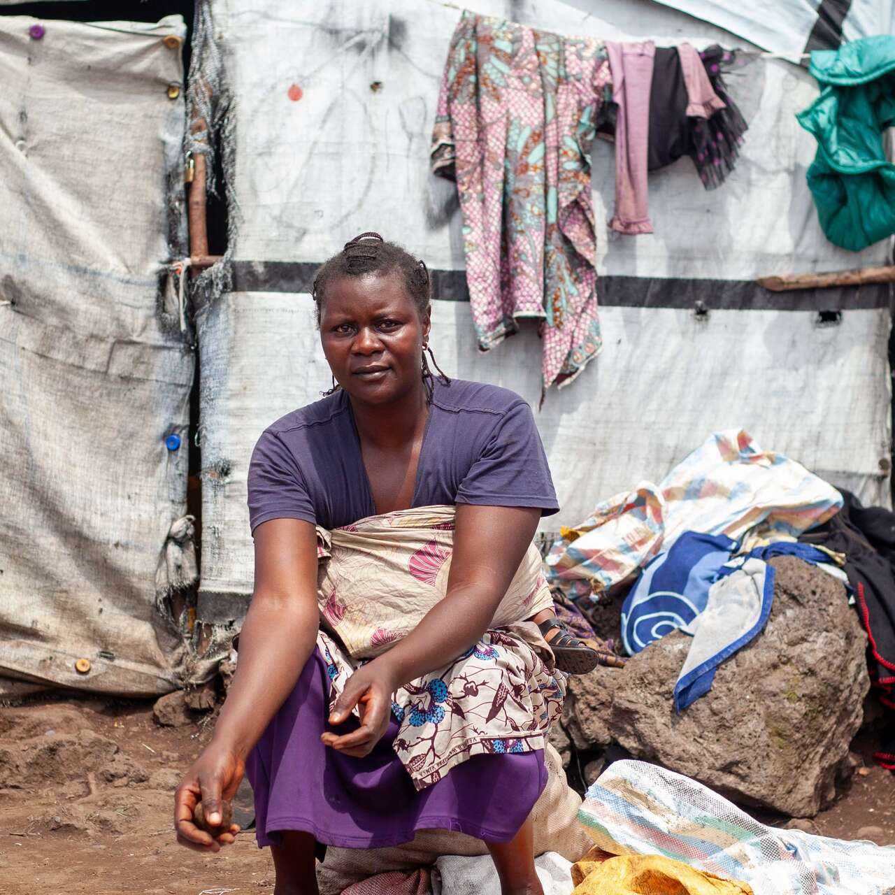 A woman sits outside, in front of a white makeshift shelter. A large pile of potatoes lie in front of her.