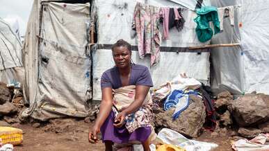 A woman sits outside, in front of a white makeshift shelter. A large pile of potatoes lie in front of her.