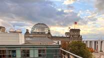 Ein Erwachsener und ein Kind stehen auf einer Terrasse mit Blick auf moderne Gebäude und einen bewölkten Himmel.