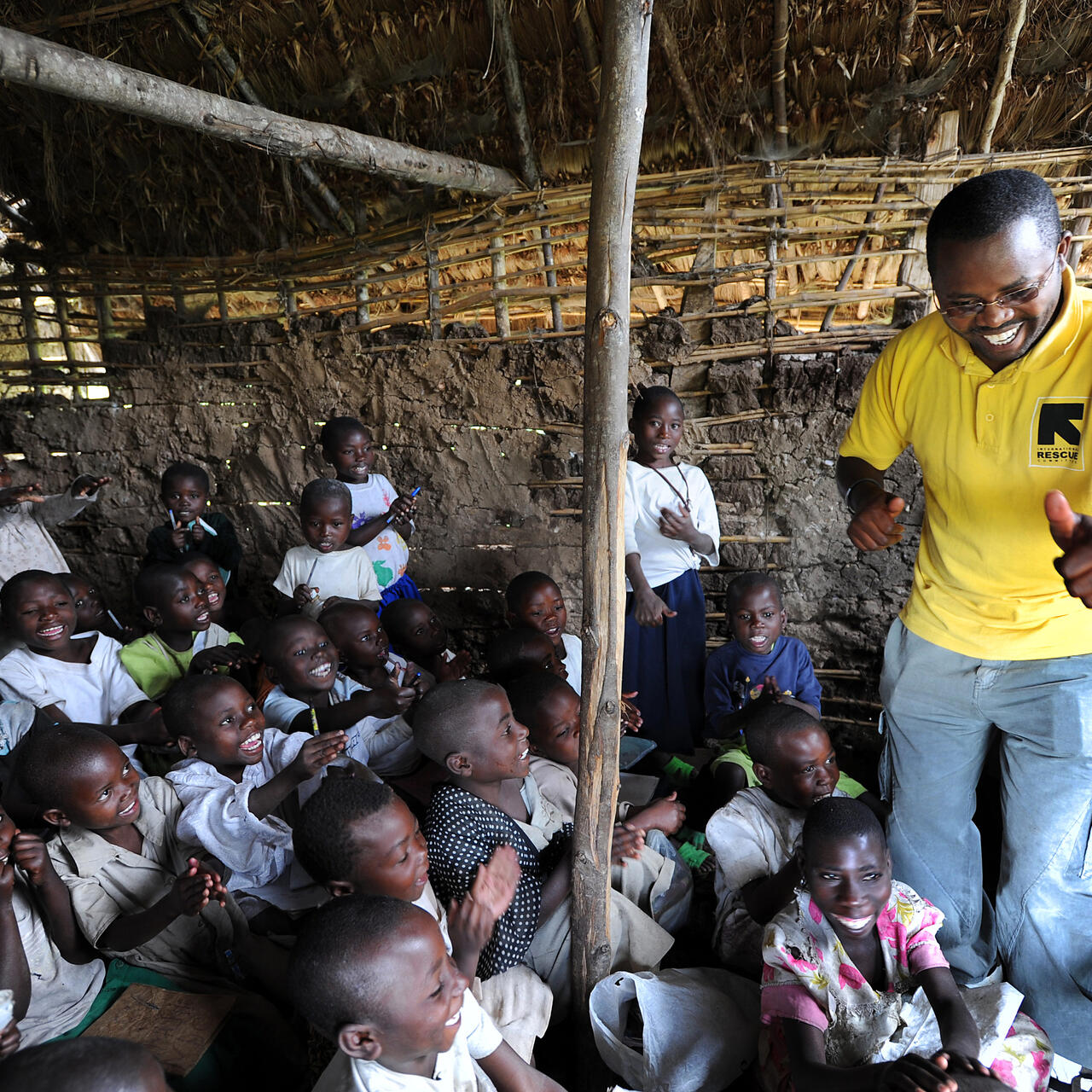 A smiling aid worker stands amidst a classroom of children, leading them in a song