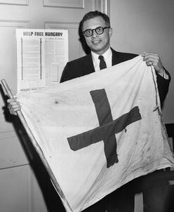 Leo Cherne poses for a picture holding a flag while standing in front of a sign that says "Help Free Hungary."