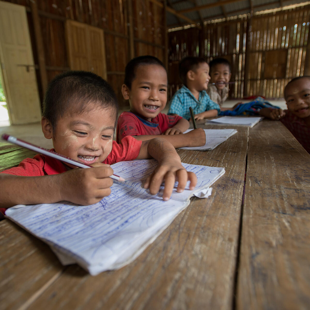 Smiling young boys in Myanmar write in their notebooks at a wooden table in tehir bamboo classroom.