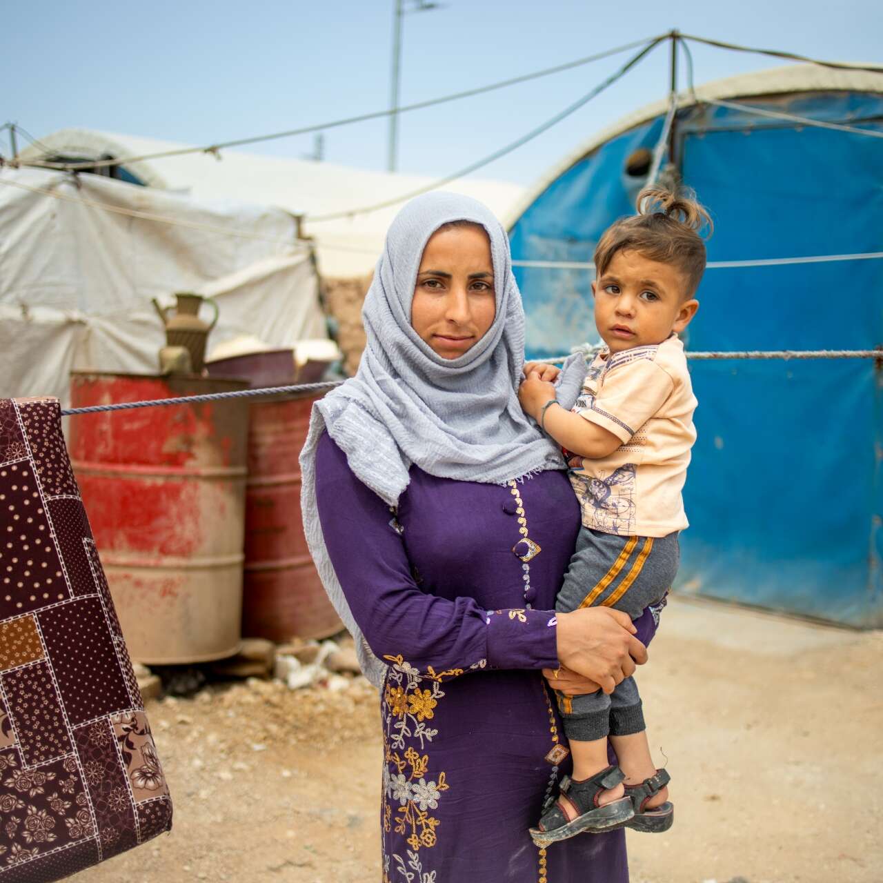 A mother holds her young son on her hip. They stand outside a make-shift shelter in a Syrian refugee camp.