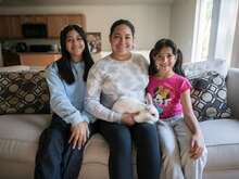 A mother sits on a couch and poses for a photo next to her two daughters. She holds the family's pet bunny in her lap.