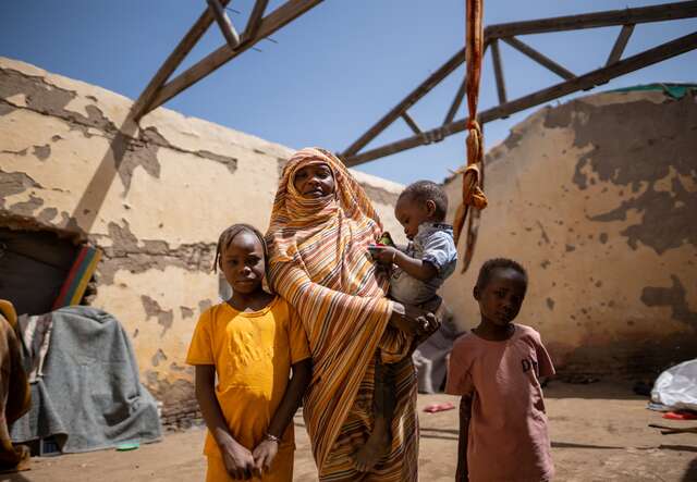 A mother holds her young child in her arms. Two other children stand by her side in a makeshift shelter in Sudan.