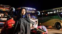 A young woman sits at a bus depot, wrapped in a blanket, next to several bags.