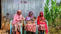 Three women sit in chairs outside and pose for a photo. The woman in the middle holds a young child in her arms.