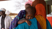 A young child clings to her mothers shoulder. The child looks towards the photographer while the mother has her backed turned.