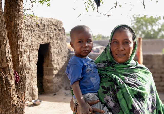 A mother holds her son on her hip outside their home in Mali. The two stare into the camera together.