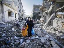 A young child wearing a yellow jacket holds an adult's hands while they walk through a destroyed neighborhood in Gaza.