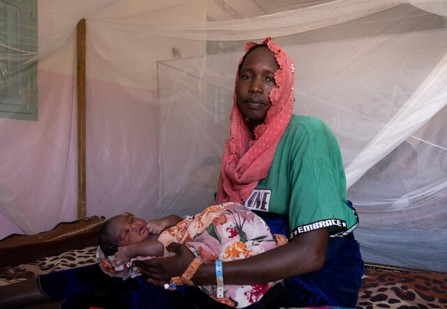 A mother sits in a hospital room with a newborn child in her lap. Mosquito netting offers a thin veil of the room behind them.