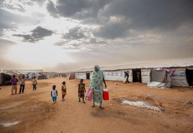 A mother walks through a camp for internally displaced people in South Sudan. She carries water while her children follow behind her.