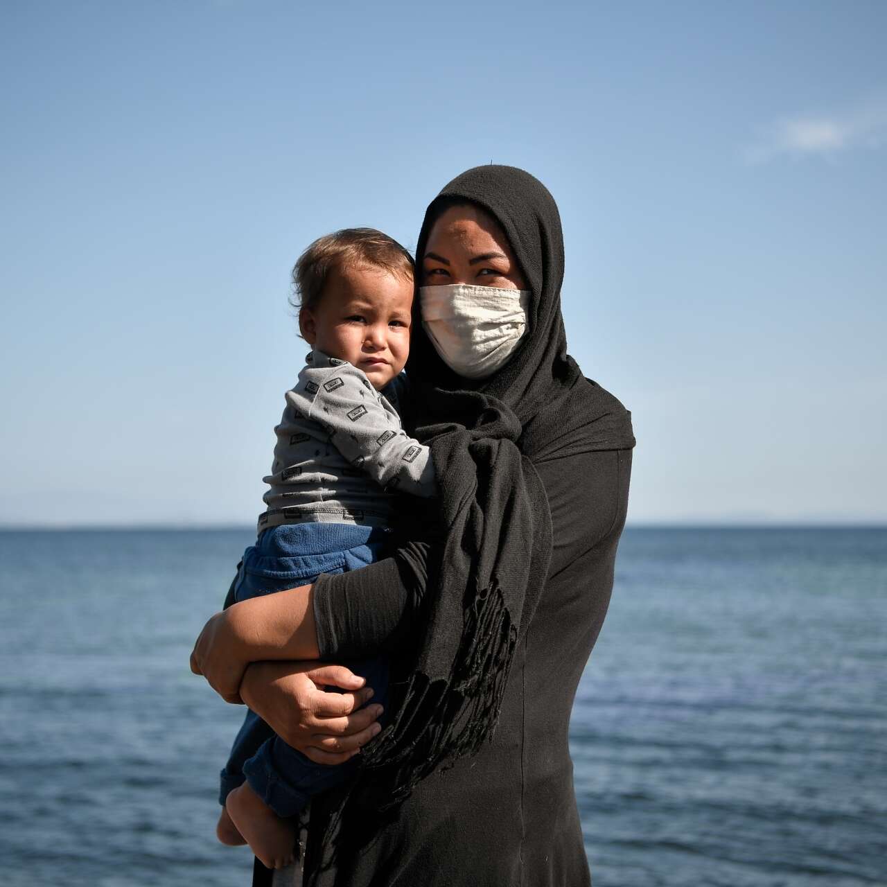 A woman holds a young child in her arms. The two are standing on the beach, in front of the ocean.