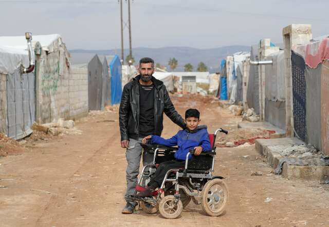 A father and his son, who is sitting in a wheelchair, pose for a photo at a camp for displaced Syrians.