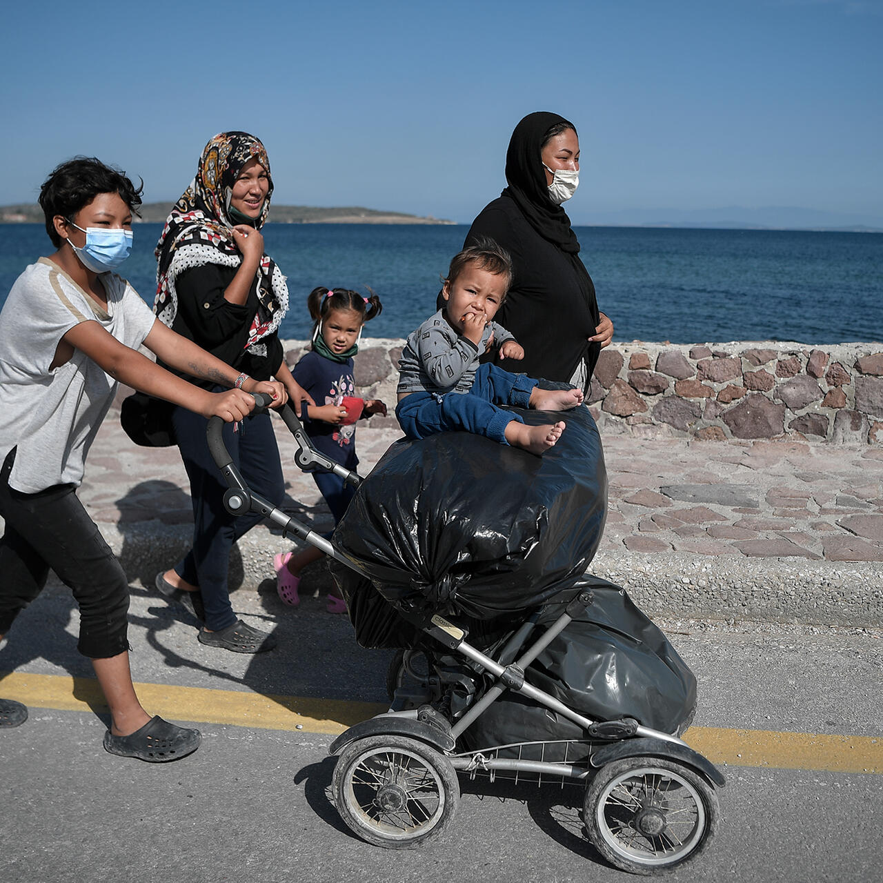 Asylum seeker mothers and their young children walk down the road in Lesvos with the sea in the background. 