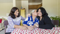 Three women, one older and two middle aged, sit at a table with a red and white table clothe. They are smiling and the older woman is kissing the woman on her left. They are all refugees. 